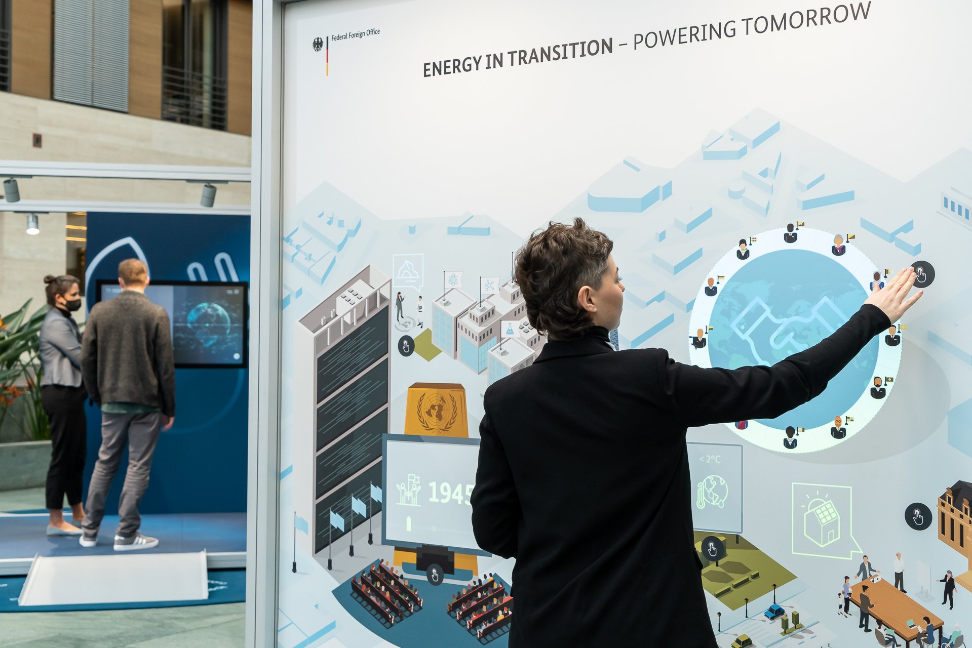 A woman stands at the Energy in Transition station of the exhibition and starts up the animation. In the background a woman and a man are standing in front of a monitor at the Renewable Energy station.